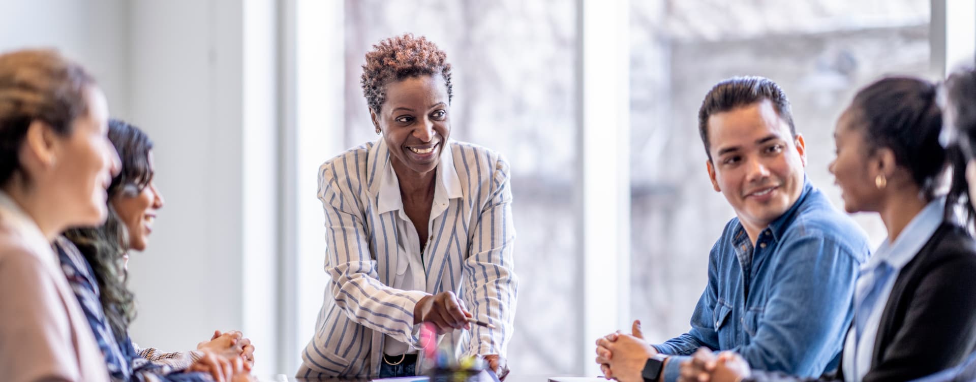 Woman in striped jacket leads an animated discussion with young team as they sit around a conference table